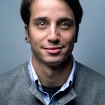 A man with short dark hair is smiling softly, wearing a light blue shirt under a gray sweater, set against a plain blue-gray background.