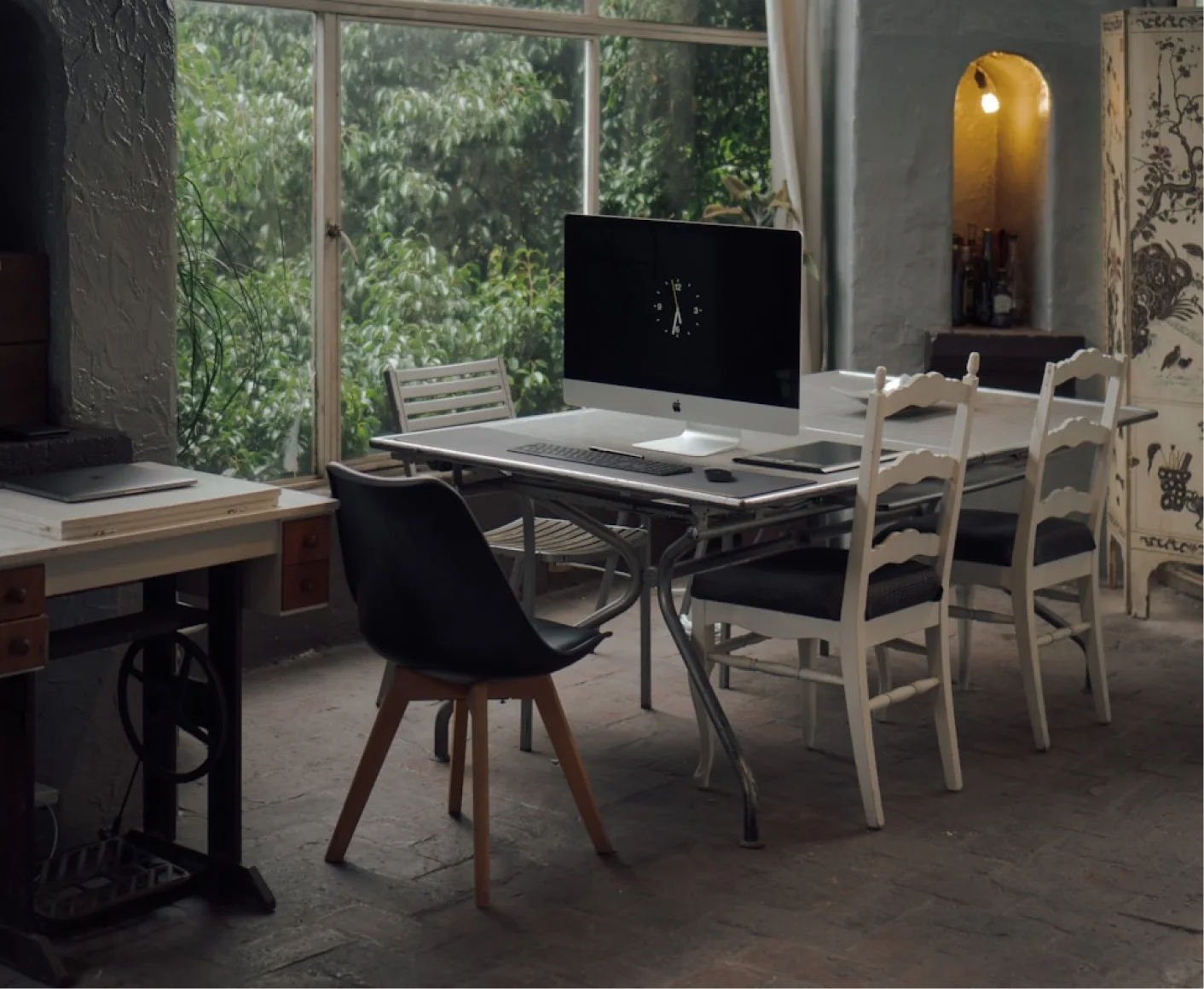 Bright architecture studio workspace with an iMac on a large table, vintage chairs, big windows overlooking green trees, and historic design elements
